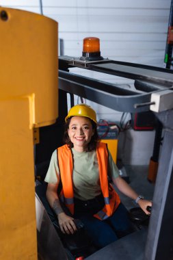 Sert şapka ve güvenlik yeleği giyen neşeli kadın forklift operatörü depodaki kameraya bakıyor, kargo...