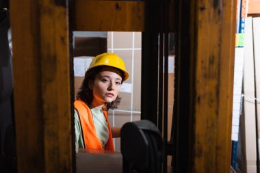 Sert şapka ve güvenlik yeleği giyen kadın forklift operatörü depodaki kameraya, kargoya bakıyor.