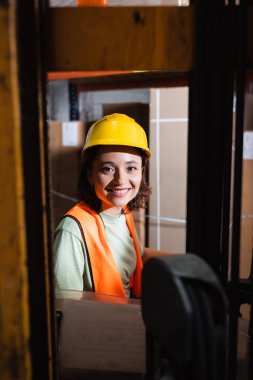Kasket ve güvenlik yeleği giyen mutlu kadın forklift operatörü depodaki kameraya bakıyor, kargo...