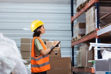 side view, female warehouse worker in hard hat and safety vest holding clipboard checking inventory clipart