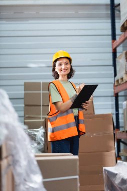 cheerful female warehouse worker in hard hat and safety vest holding clipboard checking inventory clipart