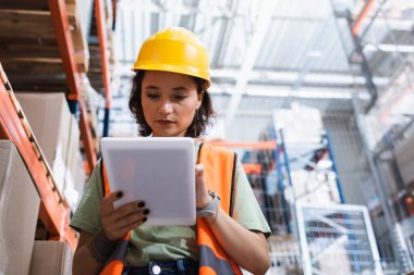 focused female warehouse worker in safety vest and hard hat holding digital tablet near cargo boxes clipart