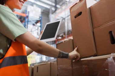 cropped female warehouse worker in safety vest smiling and holding digital tablet and checking cargo clipart