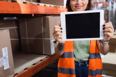 Kırpılmış ve mutlu kadın depo çalışanı güvenlik yeleği içinde elinde boş ekranlı dijital tablet tutuyor.