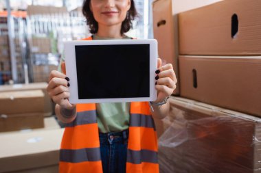 Kırpılmış ve gülümseyen kadın depo çalışanı güvenlik yeleği içinde elinde boş ekranlı dijital tablet tutuyor.