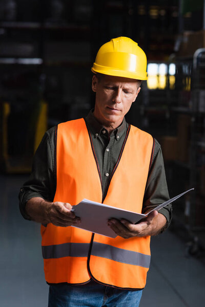 Focused middle aged supervisor in hard hat reviewing paperwork in warehouse, logistics and cargo