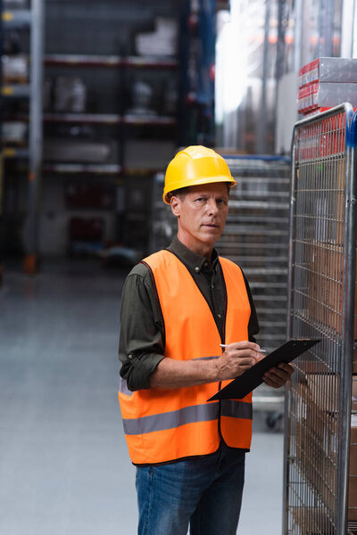 middle aged supervisor in hard hat and safety vest doing paperwork in warehouse, logistics and cargo