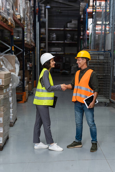 happy warehouse colleagues with clipboard and tablet shaking hands near freight on shelves