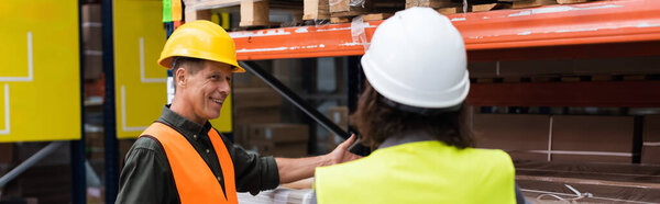 happy middle aged supervisor in hard hat gesturing and explaining work to warehouse employee, banner