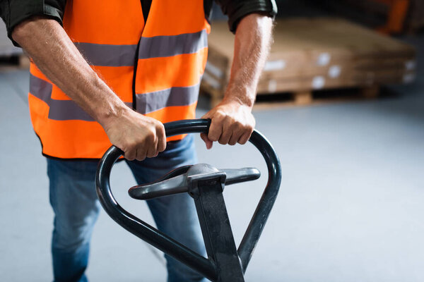 обрезанный снимок работника склада в джинсах и бронежилете, перевозящего поддон с ручной тележкой