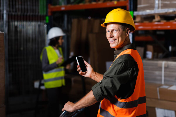 happy supervisor in safety vest using smartphone with employee in background of warehouse
