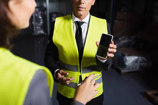cropped middle aged supervisor in safety vest using smartphone near blurred female employee