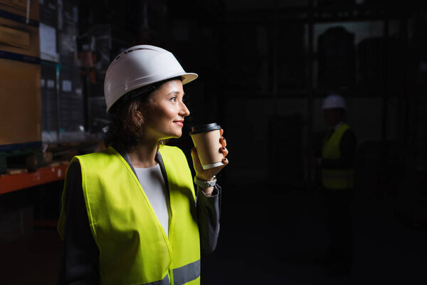 side view of woman in hard hat holding paper cup, happy warehouse employee during coffee break