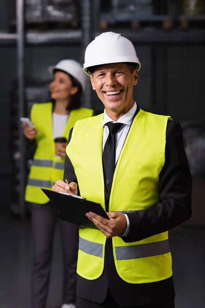 happy warehouse supervisor in hard hat holding clipboard with female employee on blurred background