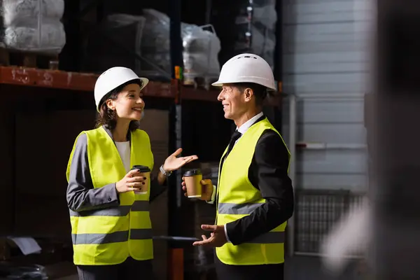 stock image logistics team, happy woman in hard hat and coffee chatting with middle aged supervisor in warehouse