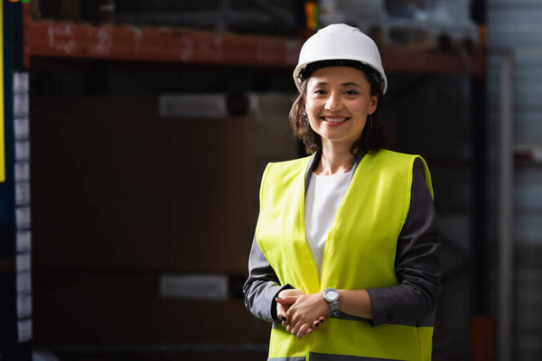 cheerful professional woman in safety vest and hard hat standing with hands in pockets in warehouse