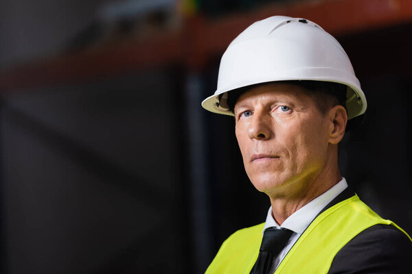 confident man in safety vest and hard hat looking at camera in warehouse, professional headshot