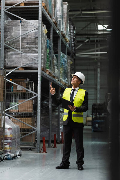 Confident supervisor in safety vest and hard hat inspecting warehouse with clipboard, professional