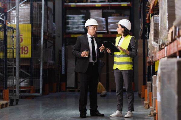 happy businesswoman in suit and heard hat holding coffee to go in warehouse, logistics management