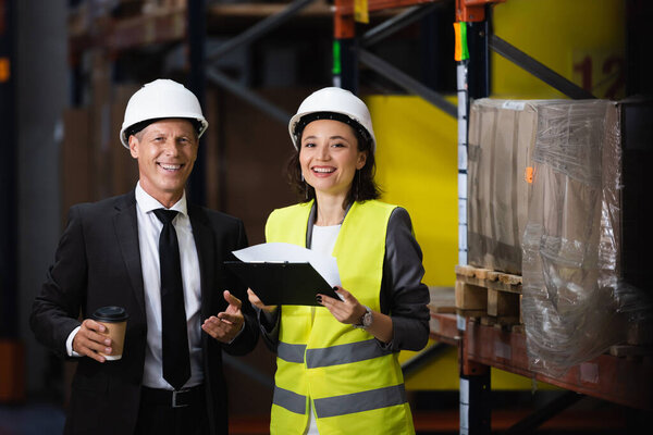 smiling businessman in suit and hard hat holding coffee near female employee, logistics concept