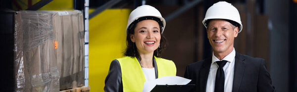 happy businessman in suit and hard hat smiling near female employee, logistics banner