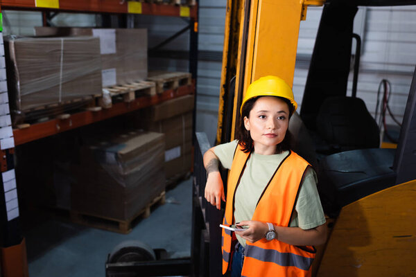 female warehouse worker in hard hat and safety vest holding smartphone near forklift, cargo