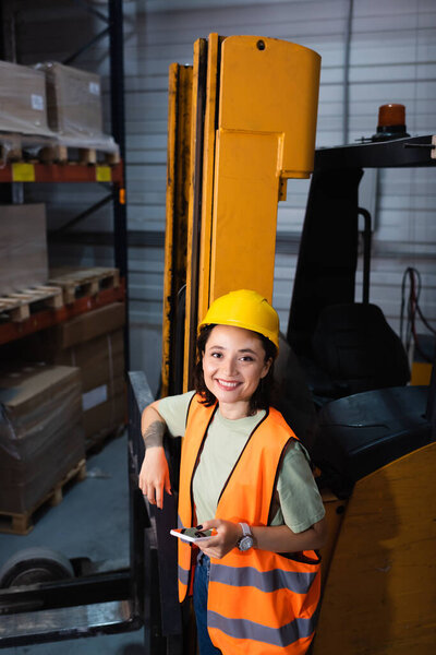happy female warehouse worker in hard hat and safety vest holding smartphone near forklift, cargo