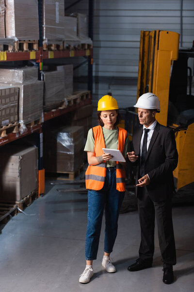 business meeting in a warehouse, supervisor in suit and hard hat near female employee with tablet