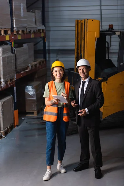 happy supervisor in suit and hard hat standing with female employee using tablet, logistic planning