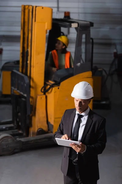 middle aged warehouse manager in business attire and hard hat using tablet while planning logistics