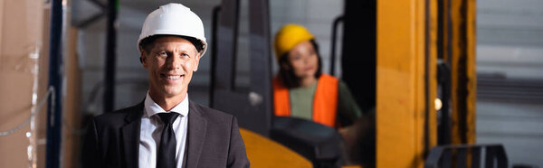 middle aged warehouse supervisor in suit and hard hat smiling while looking at camera, banner