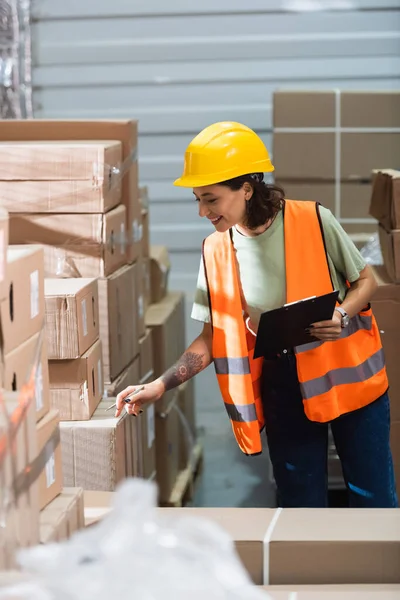 happy female warehouse worker in hard hat and safety vest holding clipboard checking inventory