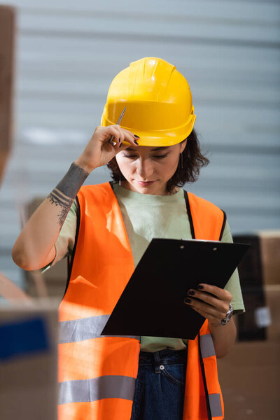 female warehouse worker in safety vest adjusting hard hat and holding clipboard and pen