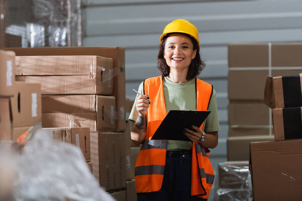 cheerful female warehouse worker in safety vest and hard hat holding clipboard and pen near cargo