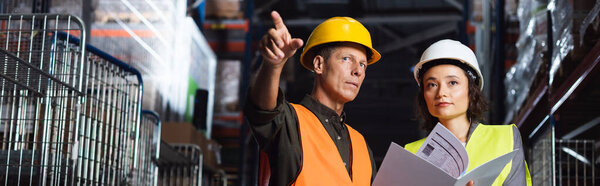 Two warehouse workers discussing logistics, middle aged man pointing near woman with folder banner