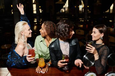 smiling multiracial fashionistas with cocktails talking during party in bar, friendship and leisure clipart