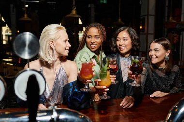 carefree multicultural girlfriends toasting with cocktails in vibrant atmosphere of night bar clipart