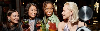 smiling and elegant multicultural female friends holding cocktails glasses in bar, horizontal banner clipart