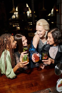 fashionable multiethnic girlfriends with cocktail glasses talking in bar, stylish atmosphere clipart