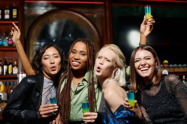 excited multiethnic girlfriends pouting lips and looking at camera in bar, glamorous lifestyle clipart