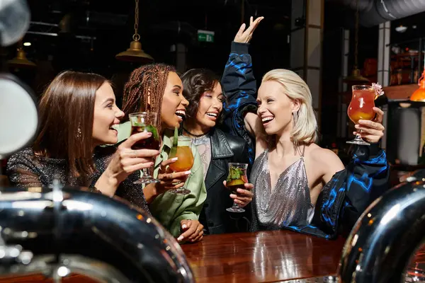 excited woman toasting with cocktail and raised hand during bar party with multiethnic girlfriends