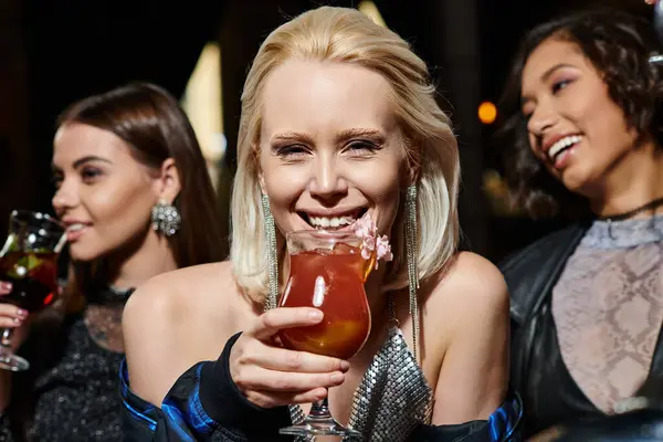 joyful blonde woman with cocktail glass looking at camera near multiethnic girlfriends in bar