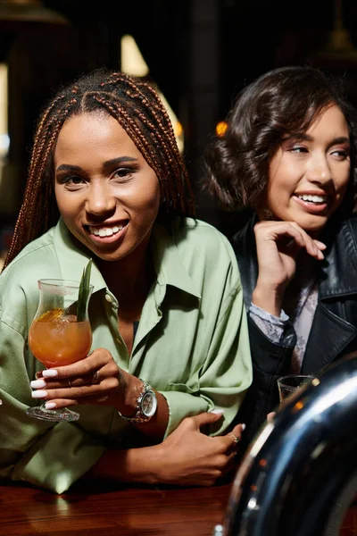 stylish african american woman with cocktail glass looking at camera near asian girlfriend in bar