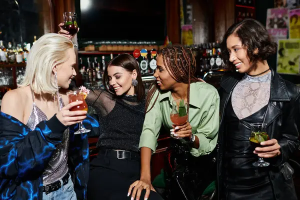 cheerful multiethnic female friends holding cocktails during conversation in modern night bar