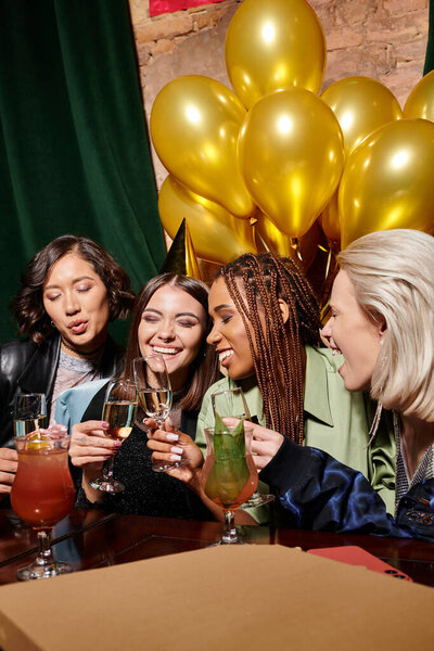 многонациональные девушки в бокалах для шампанского и закрытые глаза, смеющиеся возле золотых шариков в баре