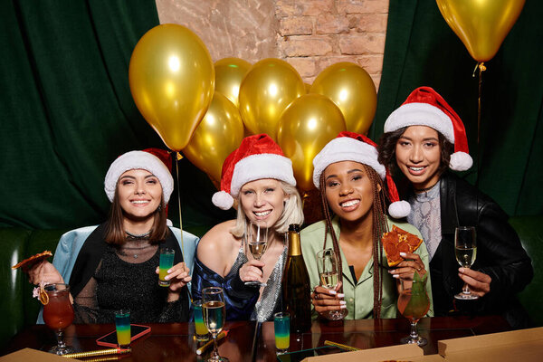 happy multiethnic women in Christmas hats looking at camera near alcohol drinks and golden balloons