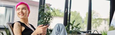 Dövmeleri olan neşeli kadın işçi. Ellerinde telefonla oturuyor. İş konsepti.