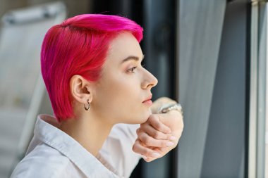 Pembe saçlı, beyaz tişörtlü genç bayan işçi hafifçe gülümsüyor ve başka tarafa bakıyor, iş konsepti.