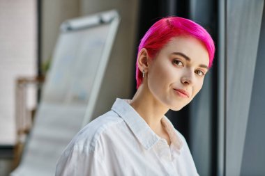 Genç pembe saçlı, küpeli, beyaz gömlekli, poz veren ve kameraya bakan bir iş kadını.