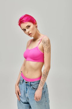 Gri arka planda poz veren pembe bluzlu, dövmeli ve kısa saçlı çekici bir kadın.